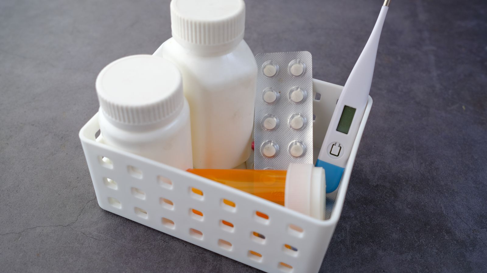 Healthcare basket with medication bottles, pills, and thermometer on a dark surface.