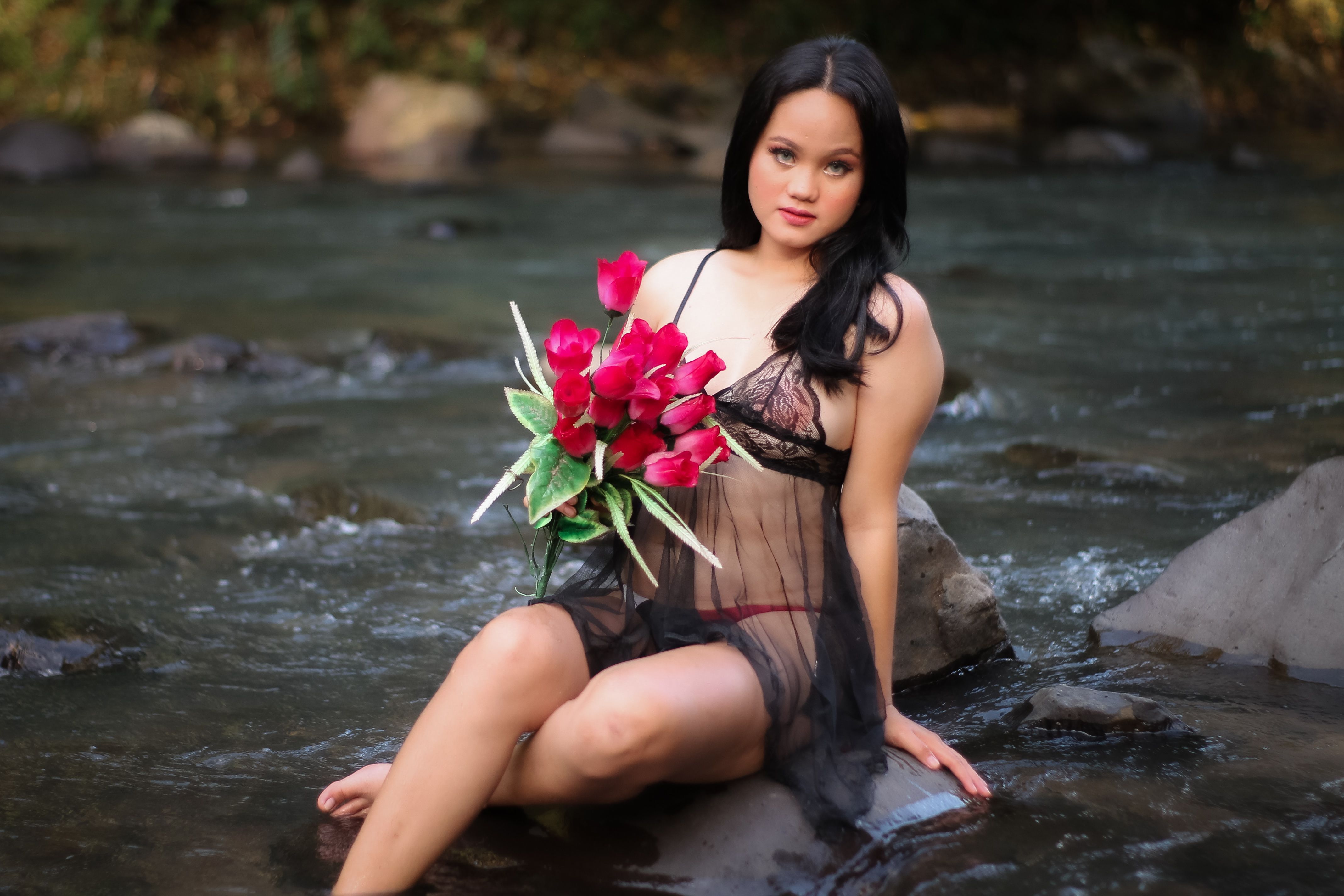 A woman in lingerie poses with a bouquet beside a tranquil river in Bulan, Philippines.