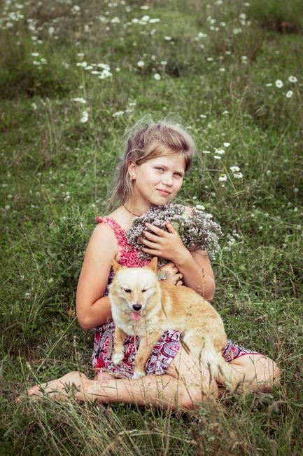 A young girl sits in a meadow holding wildflowers with a small dog by her side, capturing a serene outdoor moment.