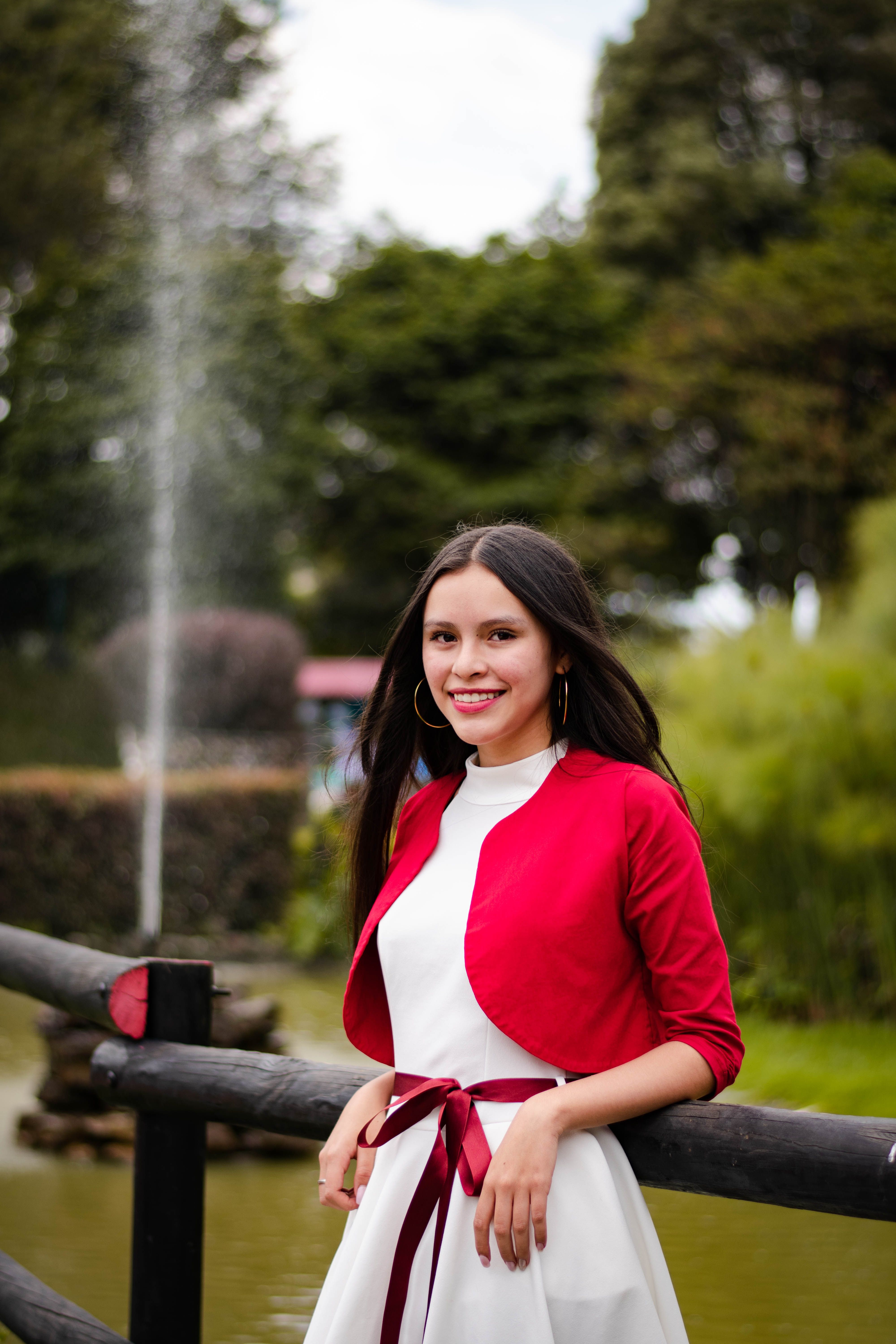 A young woman in a red jacket smiles by a tranquil fountain in a scenic park setting.