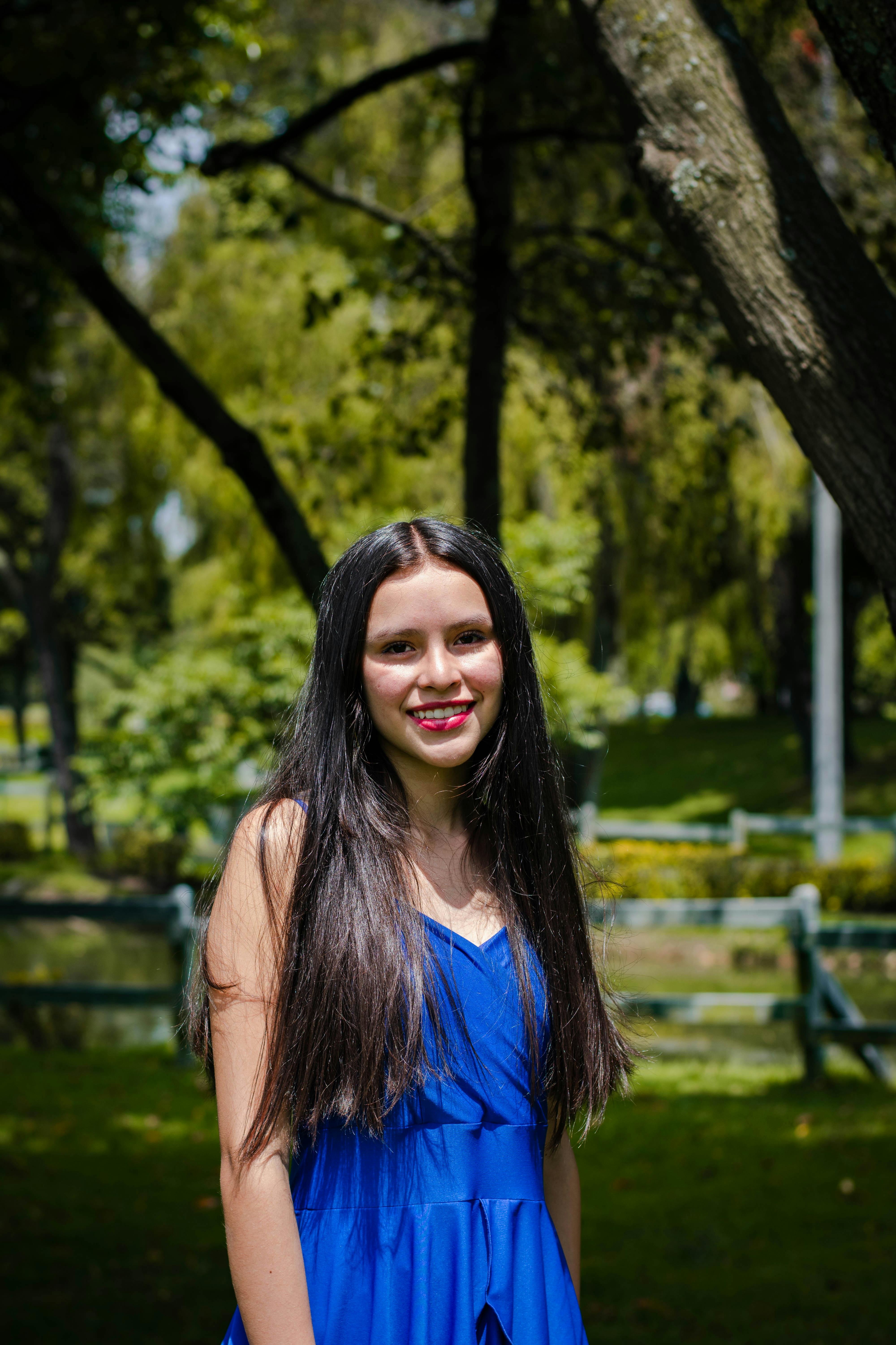 A young woman smiling in a blue dress, standing outdoors in a lush green park during the day.