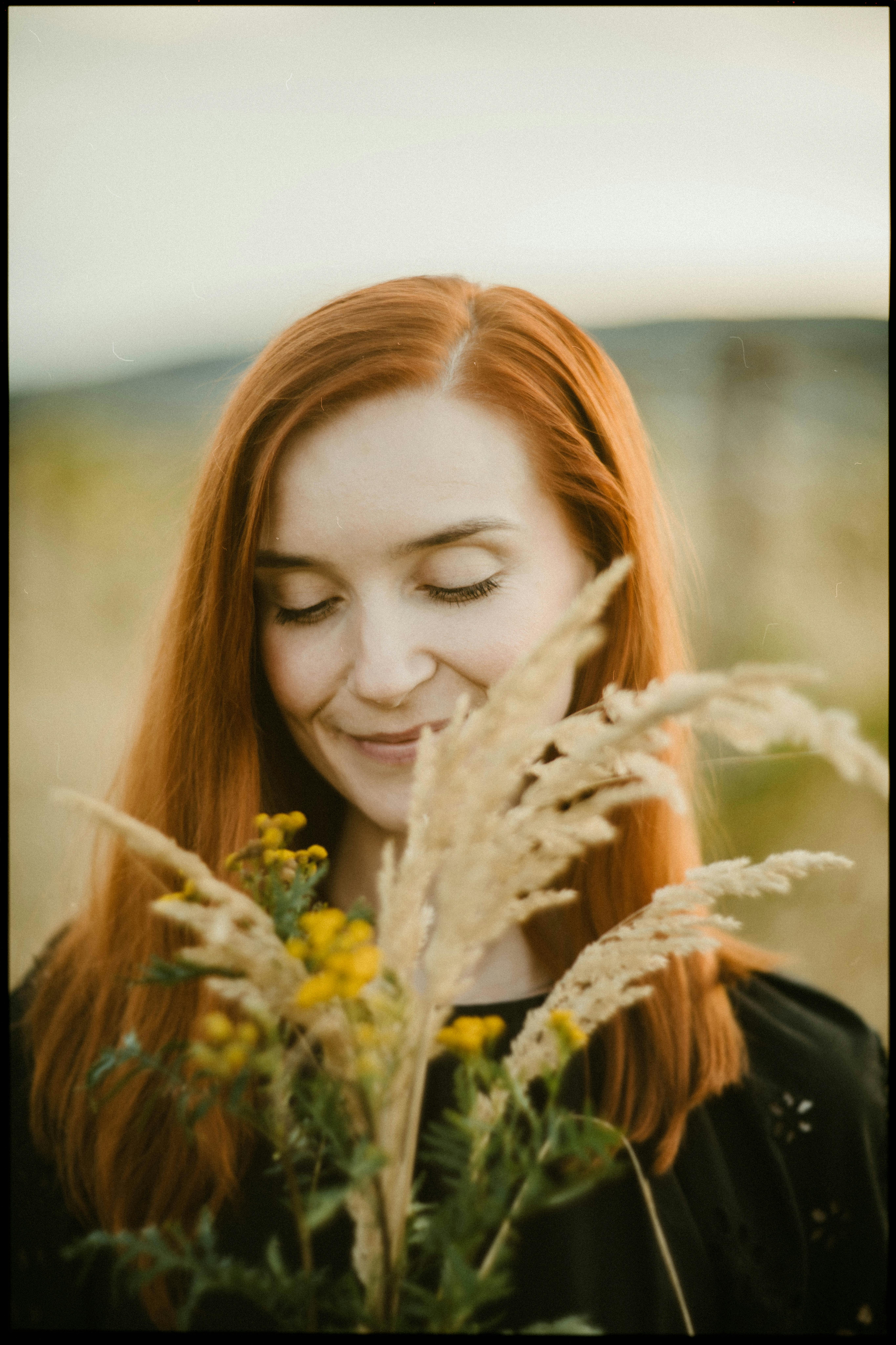 A woman with red hair enjoys a bouquet of wildflowers in a sunny field.
