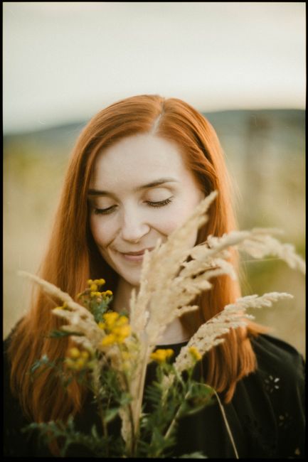 A woman with red hair enjoys a bouquet of wildflowers in a sunny field.