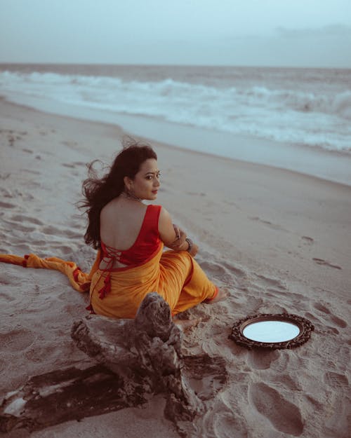 Woman in Orange Saree Sitting On Beach Sand