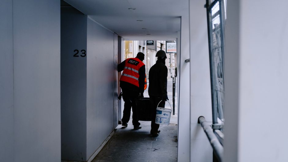 Two construction workers carrying materials in a city hallway. Two construction workers carrying materials in a city hallway.