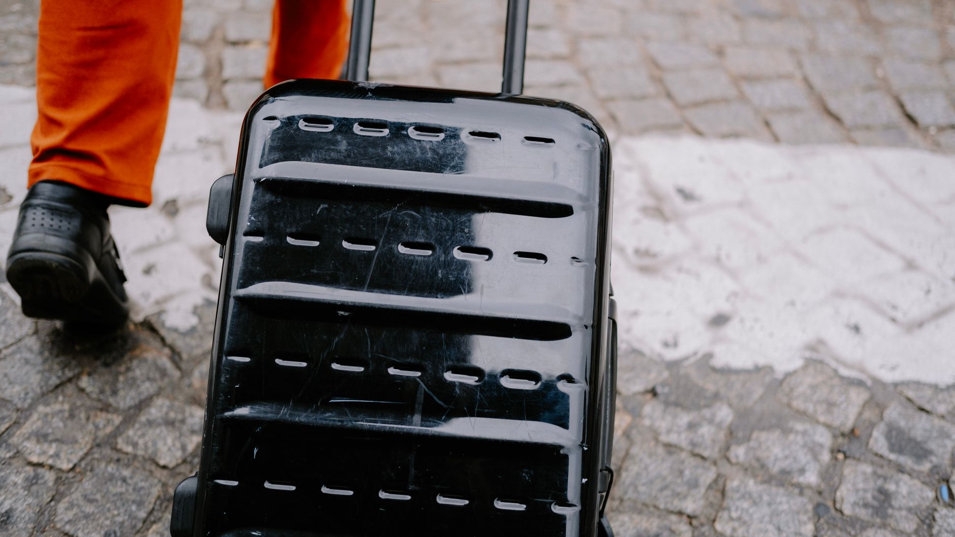 Close-up of a black suitcase being pulled on a cobblestone street by a person in orange pants.