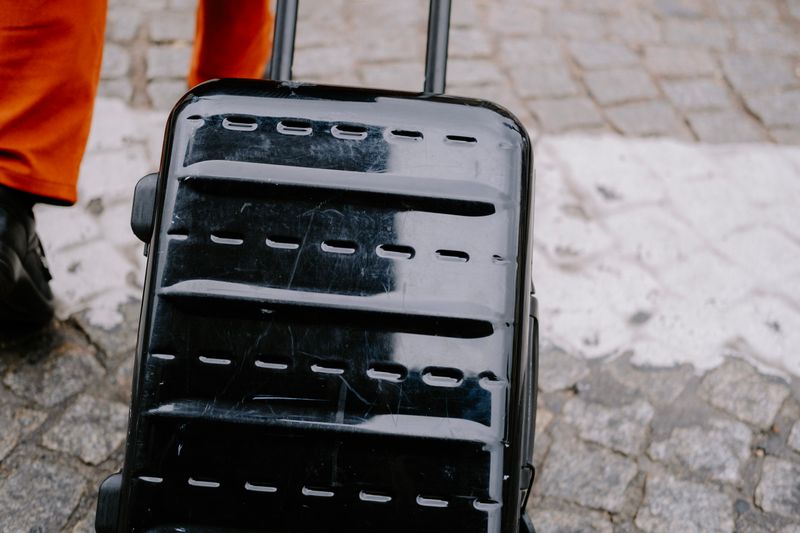 Close-up of a black suitcase being pulled on a cobblestone street by a person in orange pants.