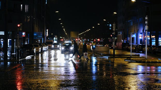 Nighttime urban street scene with reflections on wet road, pedestrians crossing and city lights.