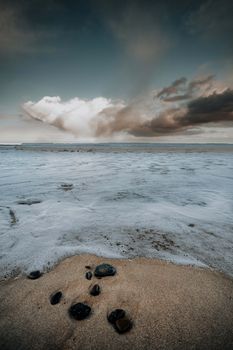 A tranquil beach scene captures rocks on sand with dramatic clouds above the ocean.