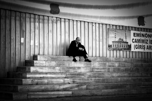 Grayscale Photo of a Man in Black Jacket Sitting on Concrete Stairs