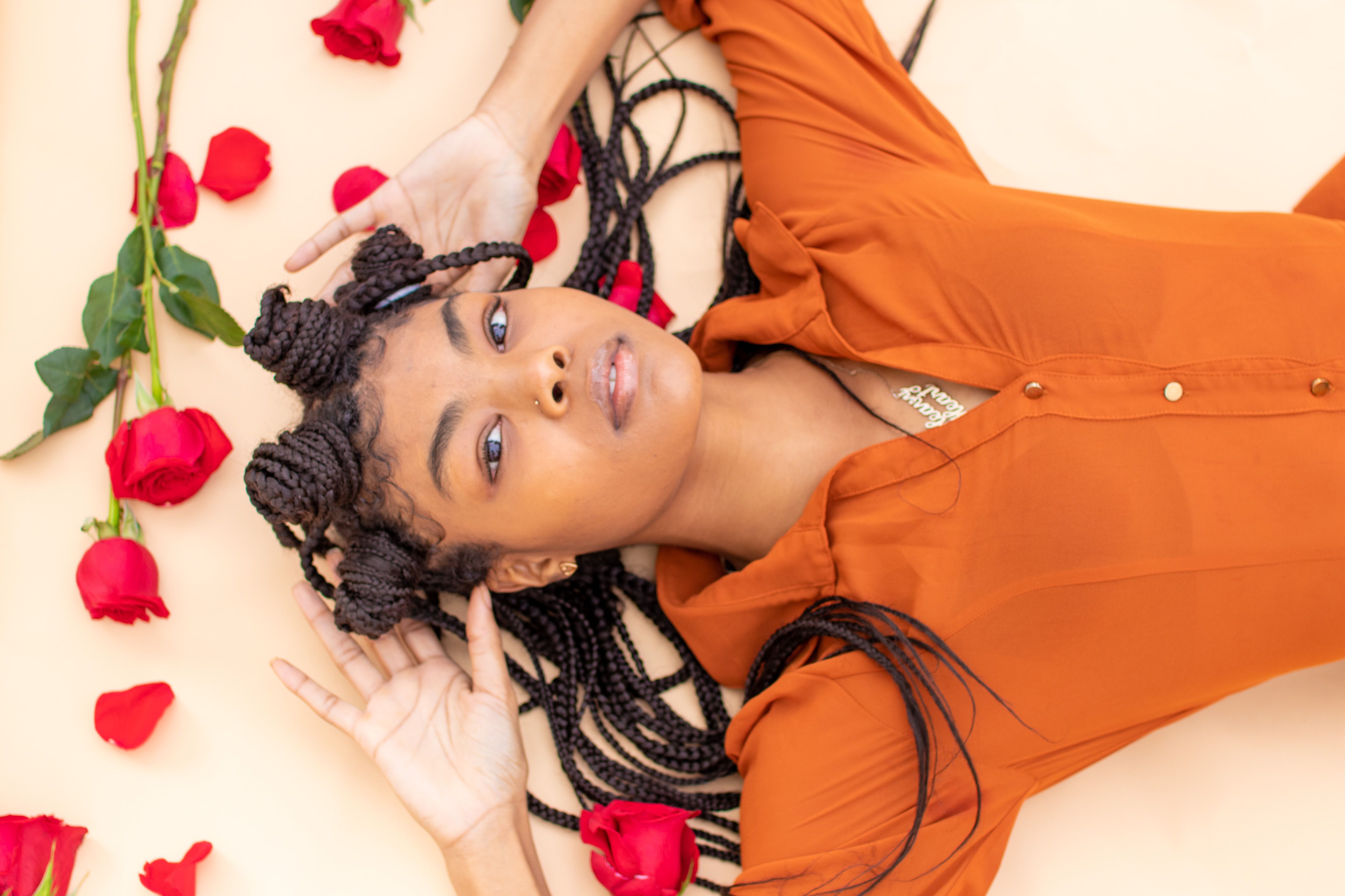 Captivating overhead portrait of a woman with braided hair surrounded by red roses, wearing a vibrant orange shirt.