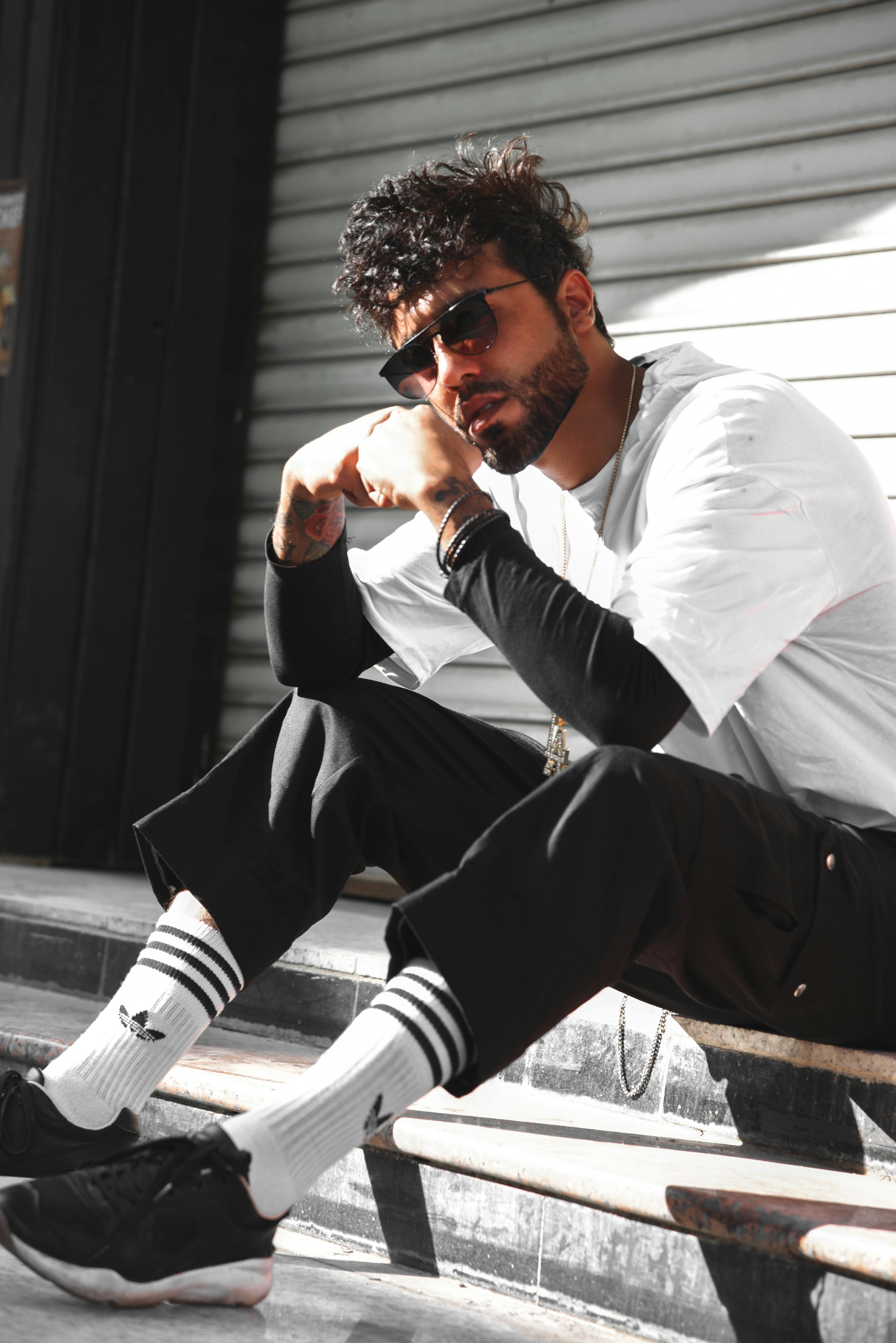 Free Stylish man posing in casual streetwear sitting on a sidewalk in urban cityscape. Stock Photo