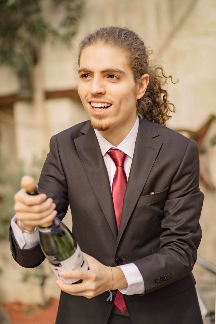 Long Haired Man In Black Suit Opening A Bottle Of Champagne 