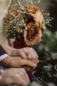 Close-up of a bride and groom holding hands with a bouquet in the background.