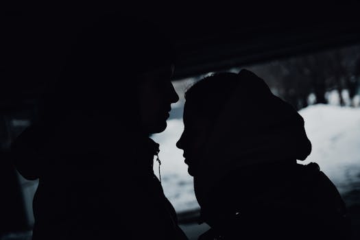 A couple in winter clothing share an intimate moment with a silhouette effect against a snowy backdrop.