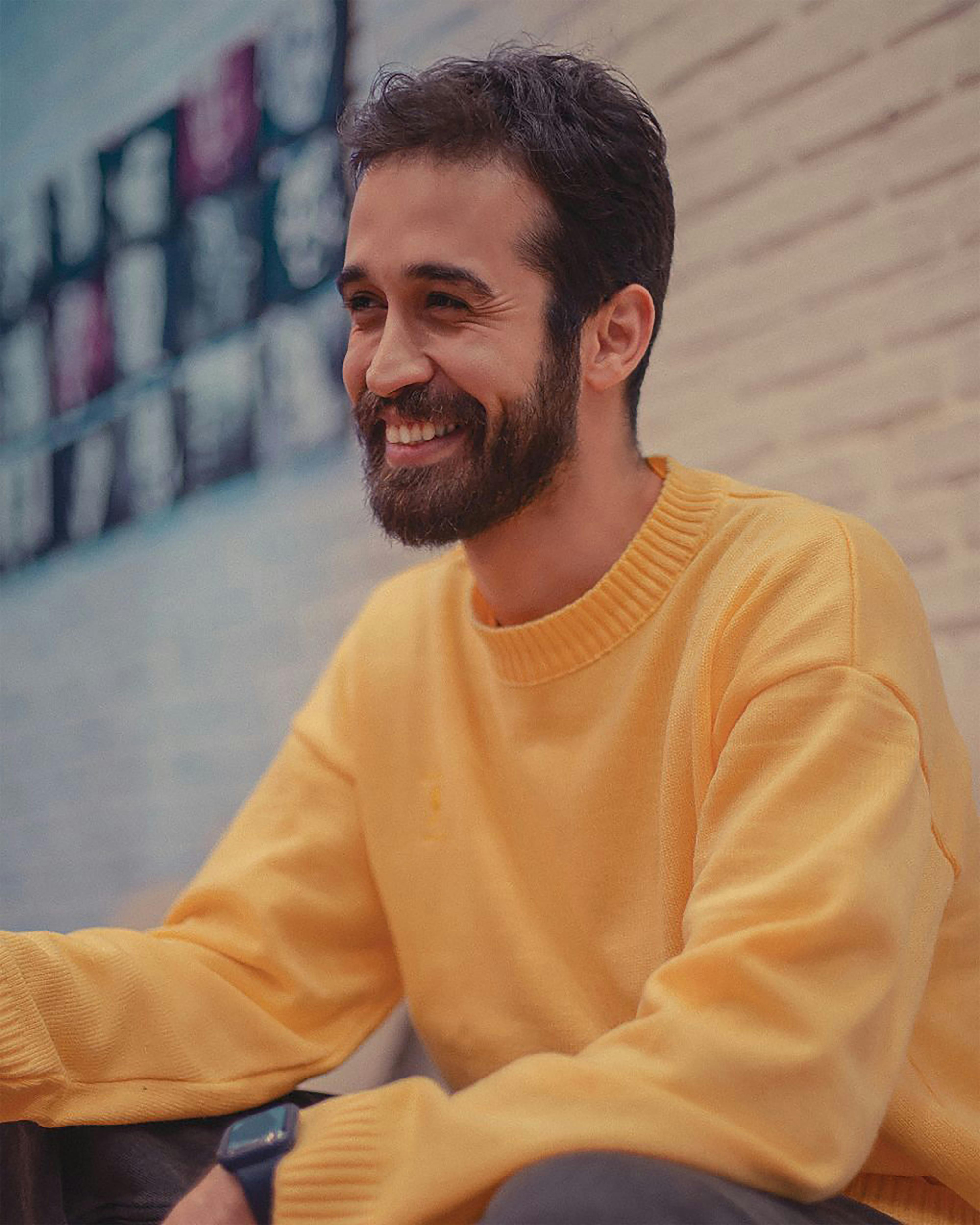 Man with beard smiling warmly in an indoor setting, wearing a yellow sweater.