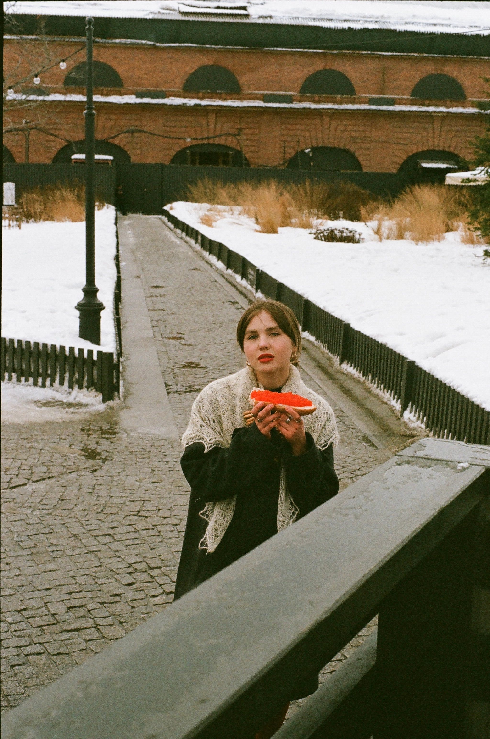 Caucasian woman standing outdoors in winter holding bread with red caviar, showcasing a snowy street scene.