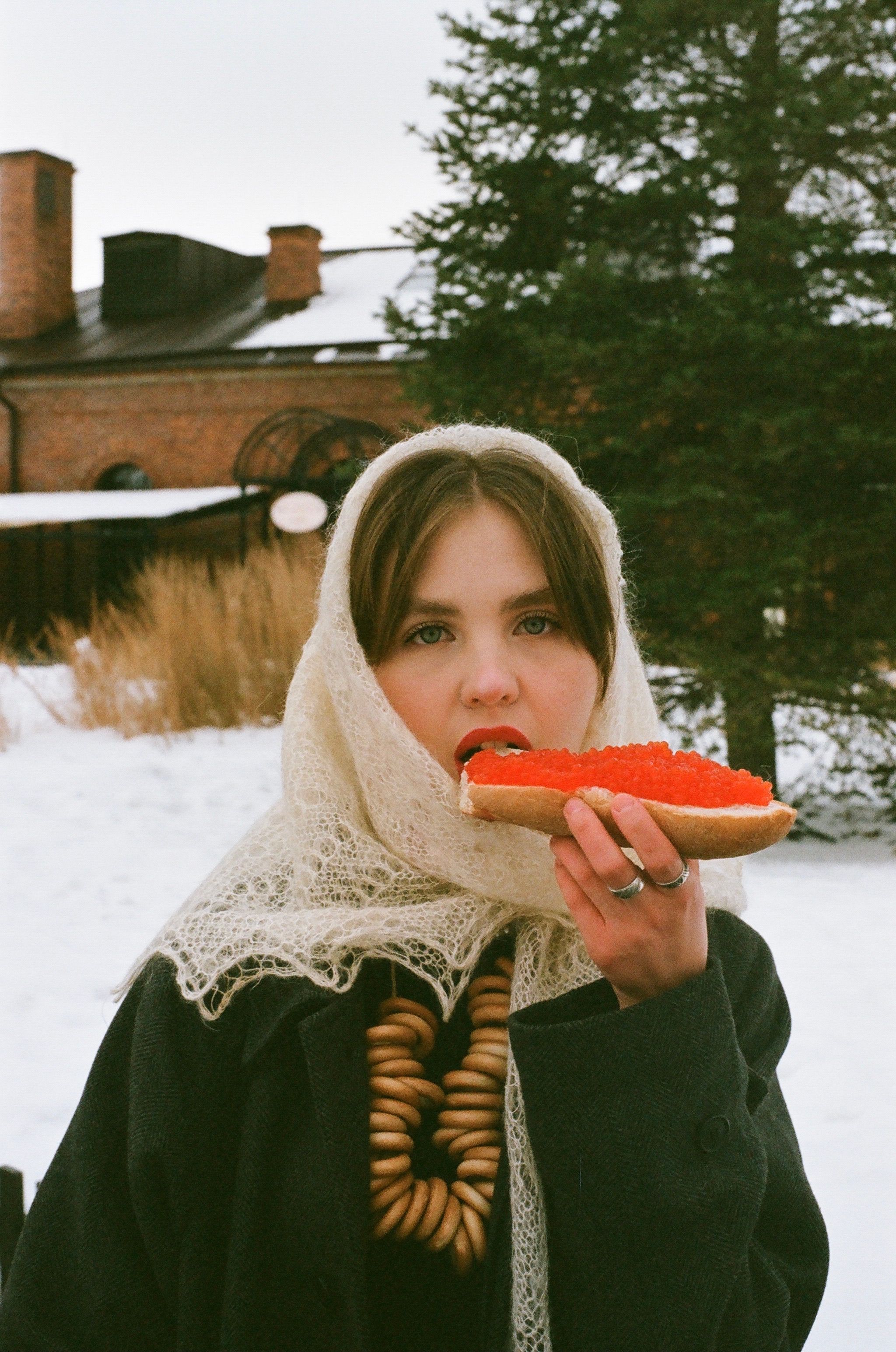 A young woman with brown hair enjoys a meal outdoors in winter attire, wearing a headscarf.