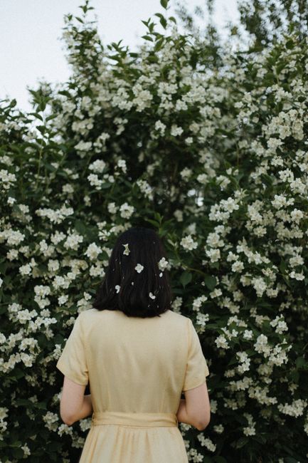 A woman with black hair stands amongst blooming white flowers, wearing a yellow dress.
