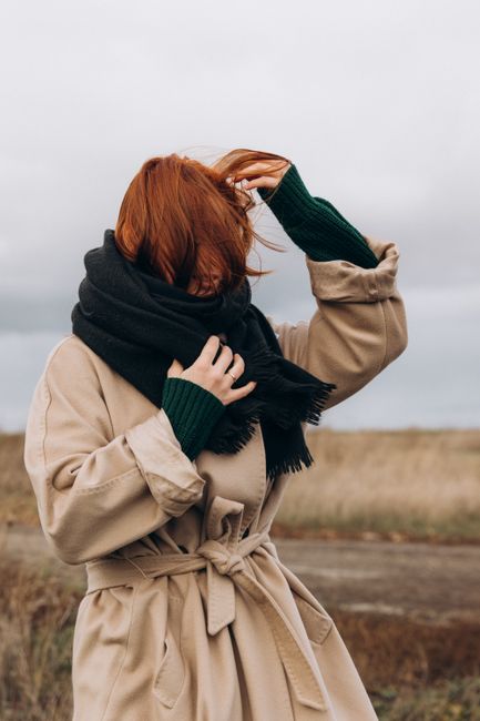 A woman with red hair in a beige coat and dark scarf stands outdoors, obscuring her face with her hand.
