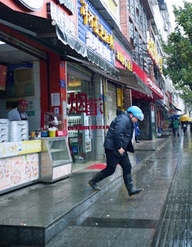 A man in a blue helmet walks quickly on a wet sidewalk on a rainy day.