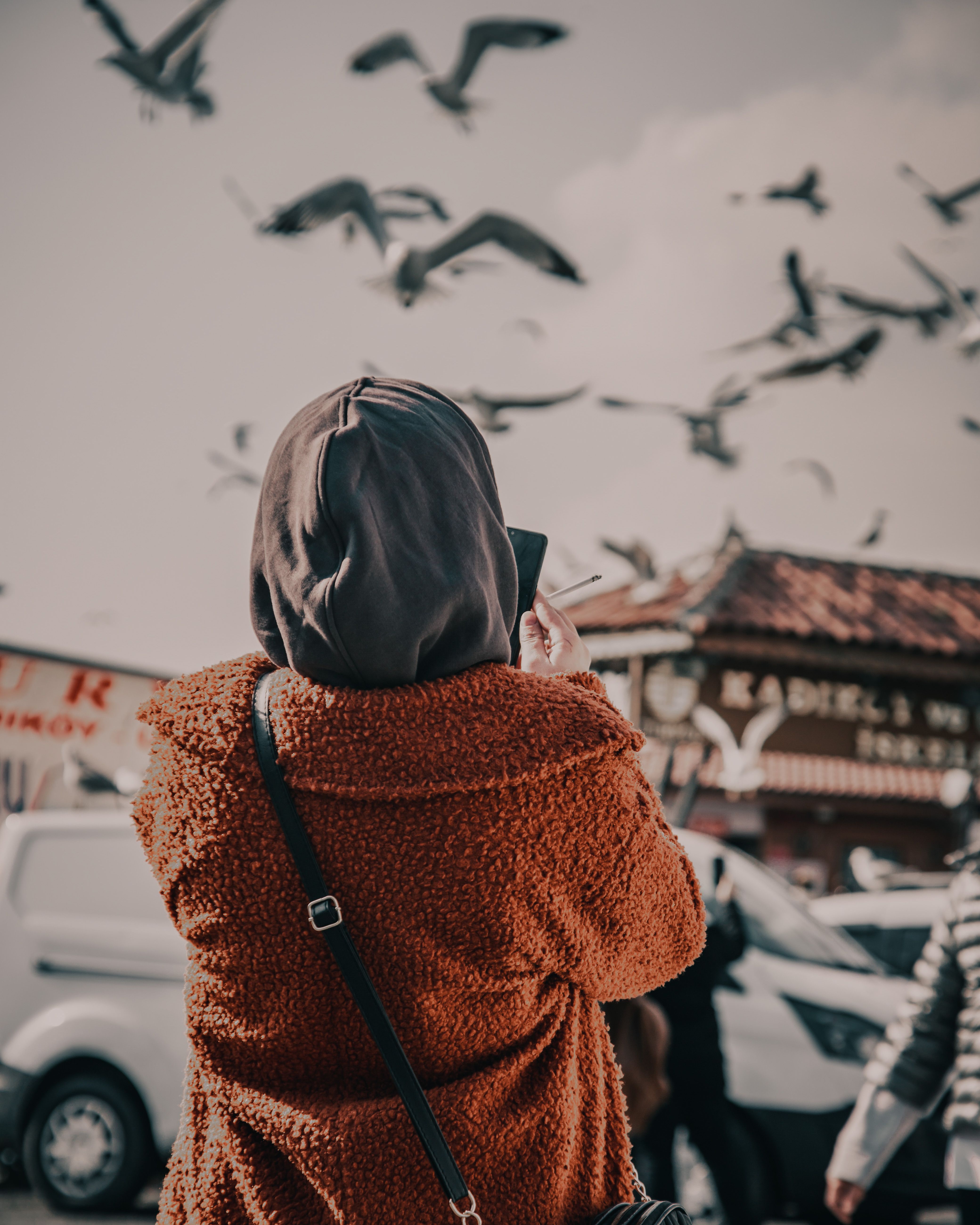 A woman in a coat takes pictures of birds flying high in an urban setting.