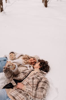 Smiling couple in winter clothes lying on snow, enjoying the outdoors.