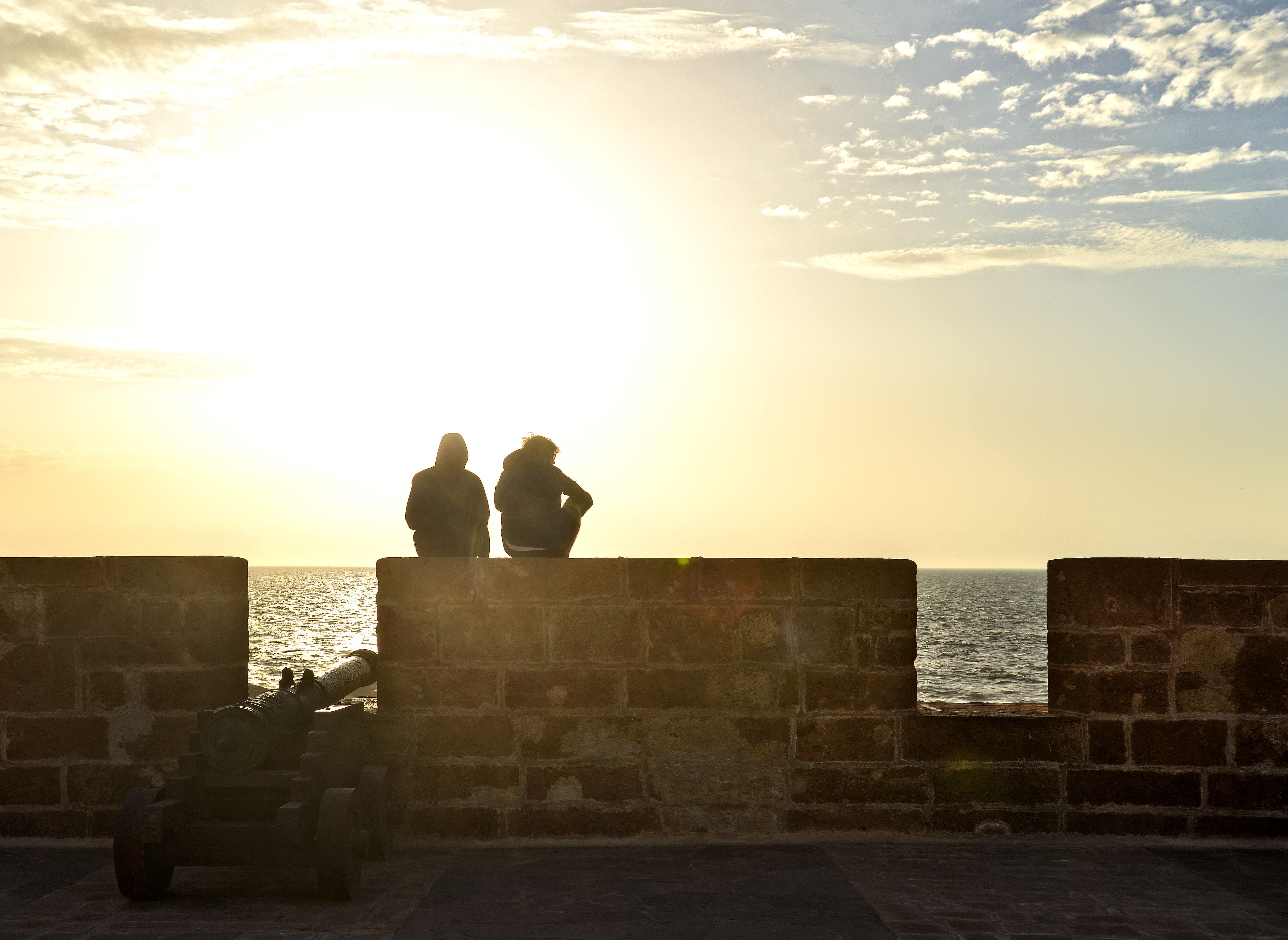 Two people silhouette sitting on a wall by the ocean during sunset in Morocco.