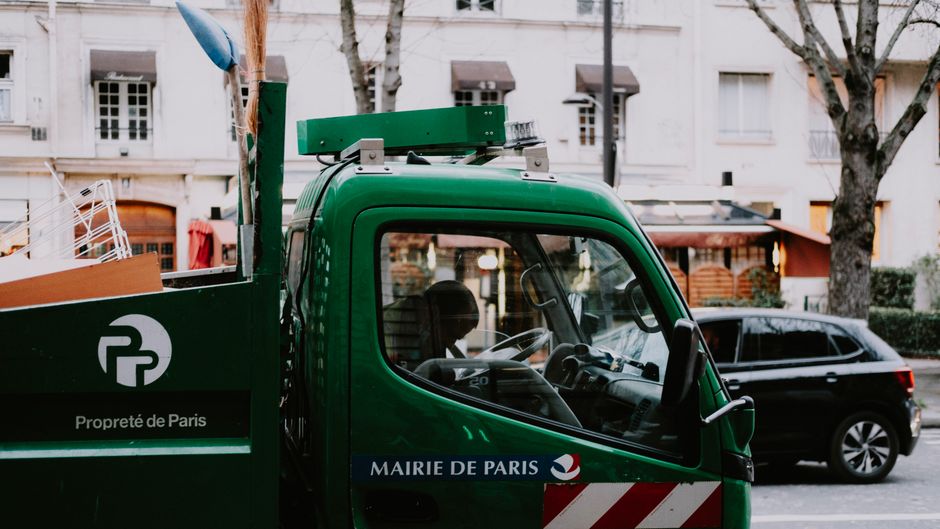 A green city garbage truck, marked 'Propreté de Paris,' on a Parisian street with urban backdrop.