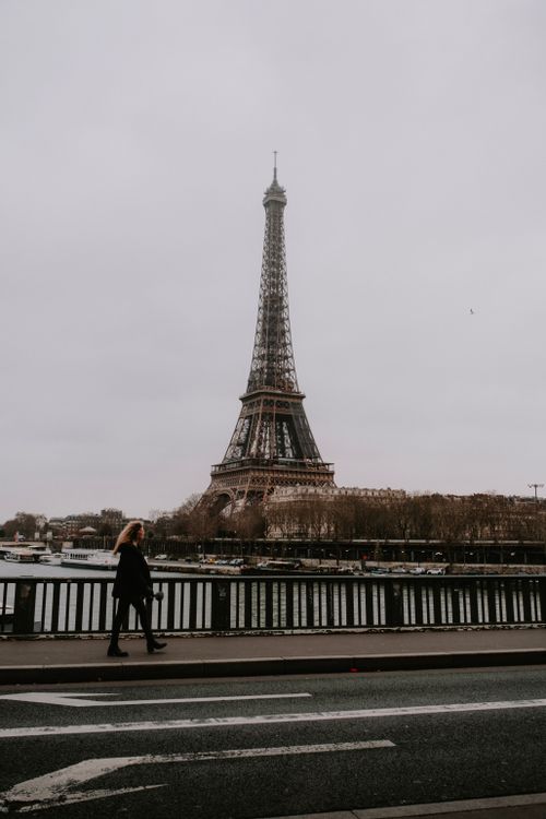 Free A woman walks on a Parisian bridge with a view of the Eiffel Tower on an overcast day. Stock Photo