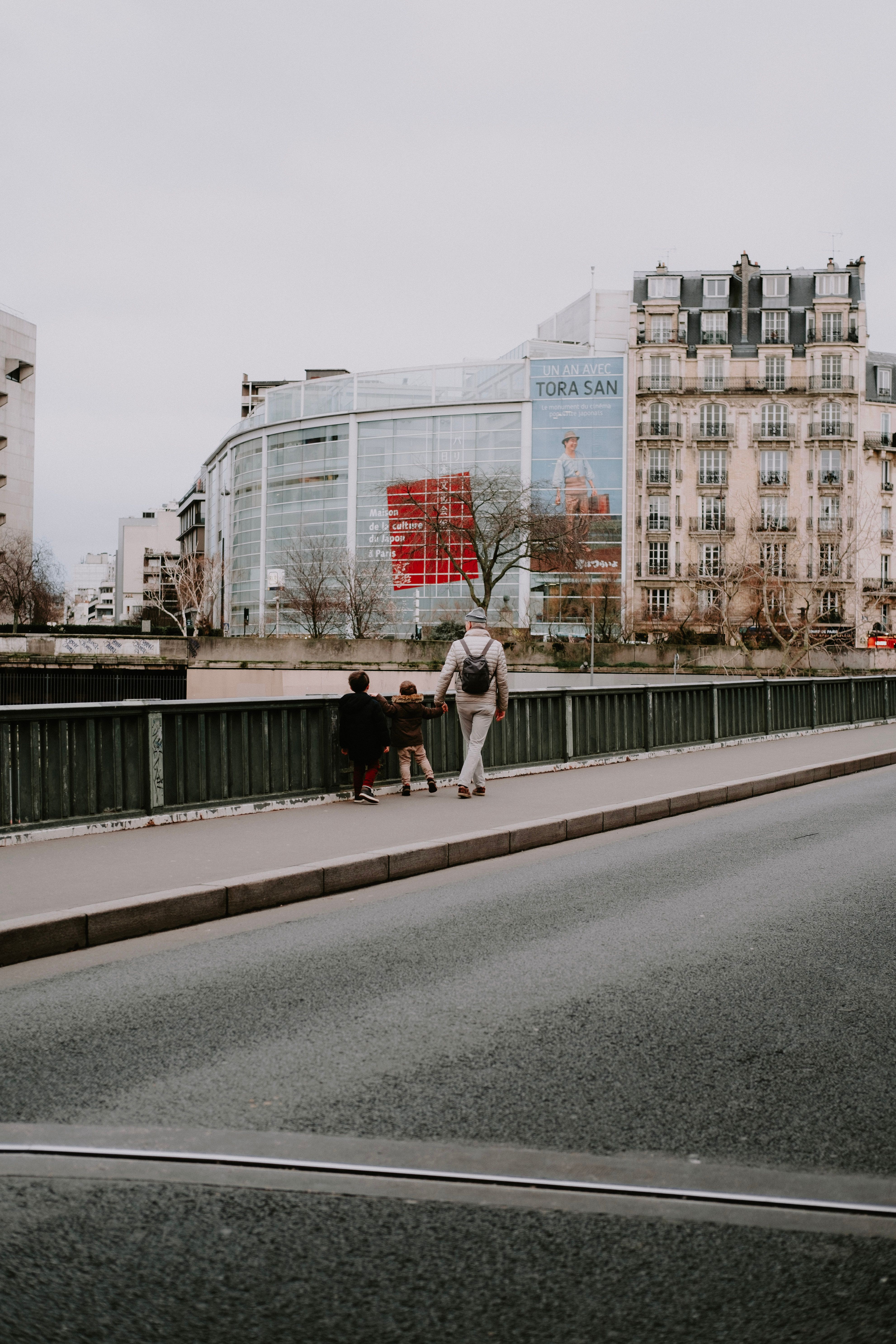 City scene with people walking along a bridge, featuring urban architecture and a prominent billboard.