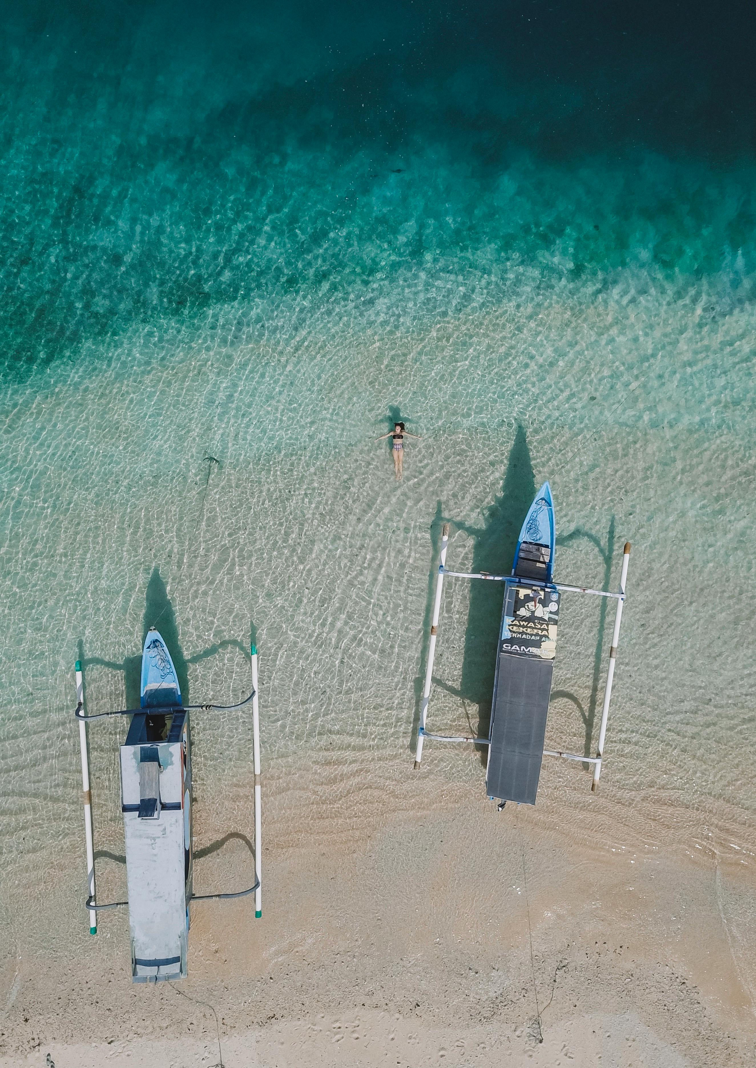 Aerial shot showcasing two boats and a woman swimming on a tropical beach coastline.