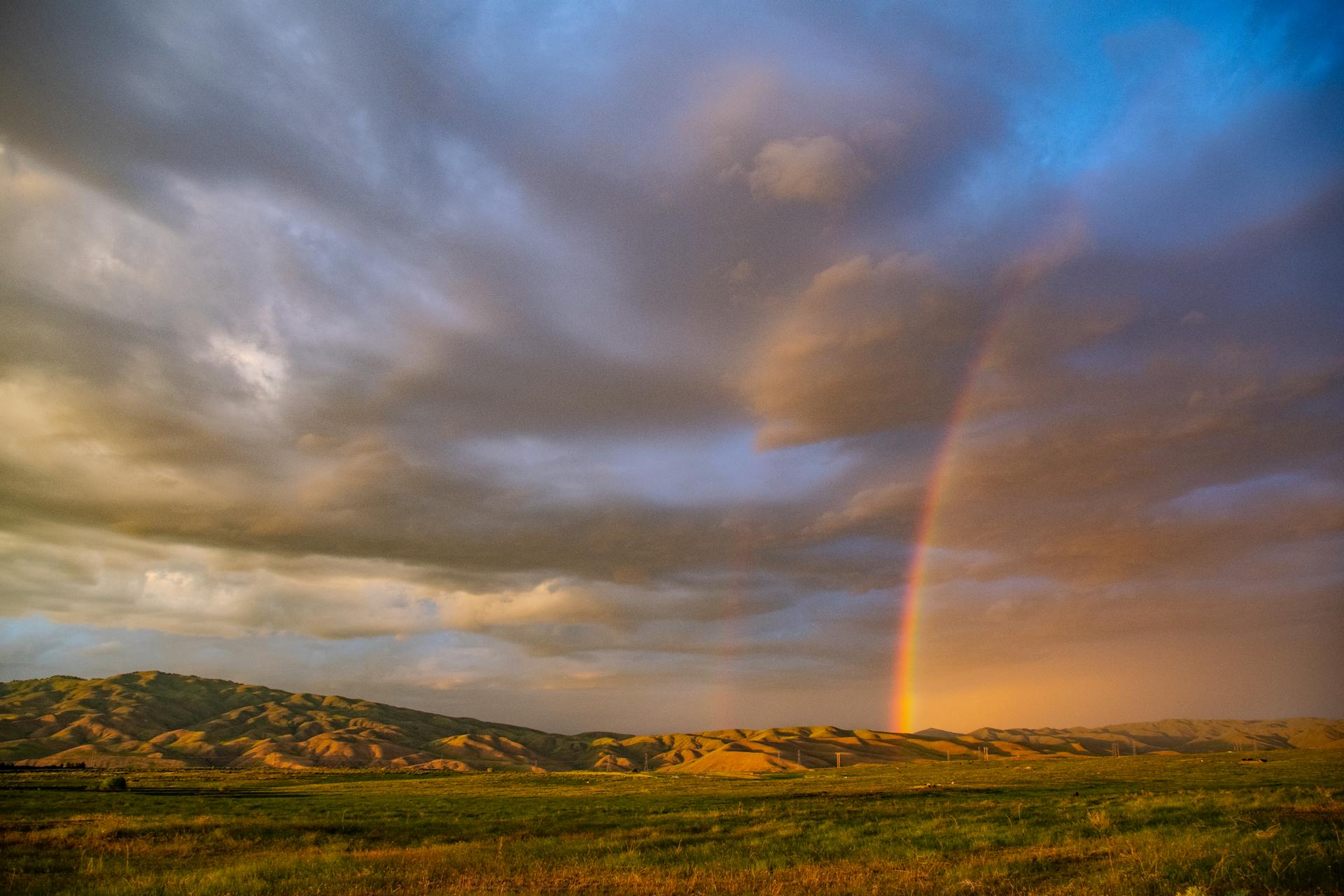 Dramatic sky with a rainbow over a vast scenic field and mountains.