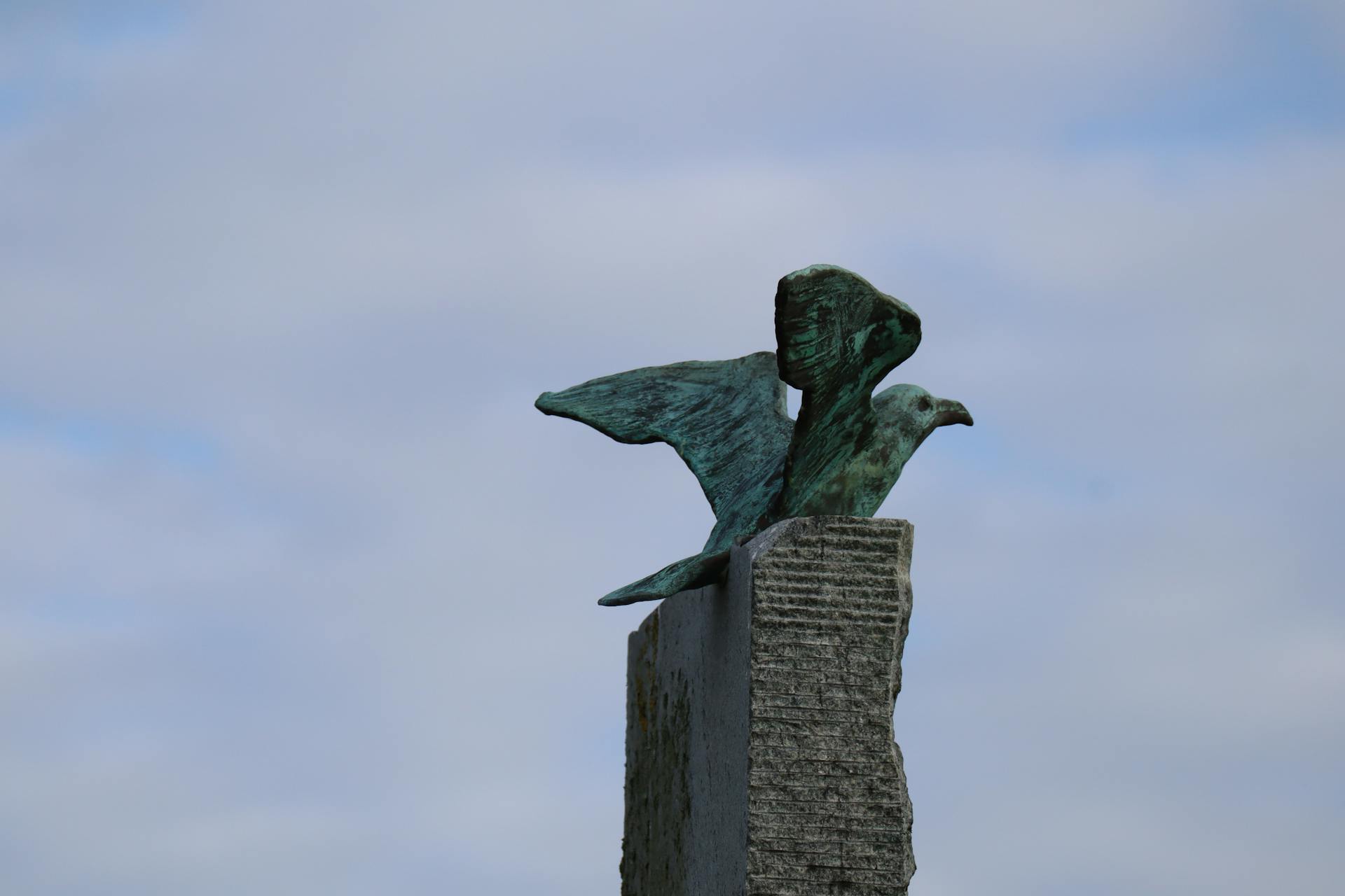 Close-up of a bronze bluebird sculpture on a stone pedestal against a cloudy sky.