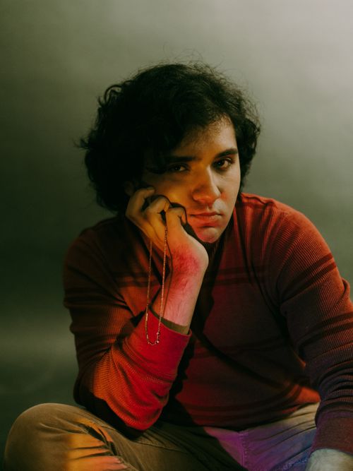 Free Portrait of a young man sitting with hand on cheek, dimly lit with warm tones against a gray background. Stock Photo