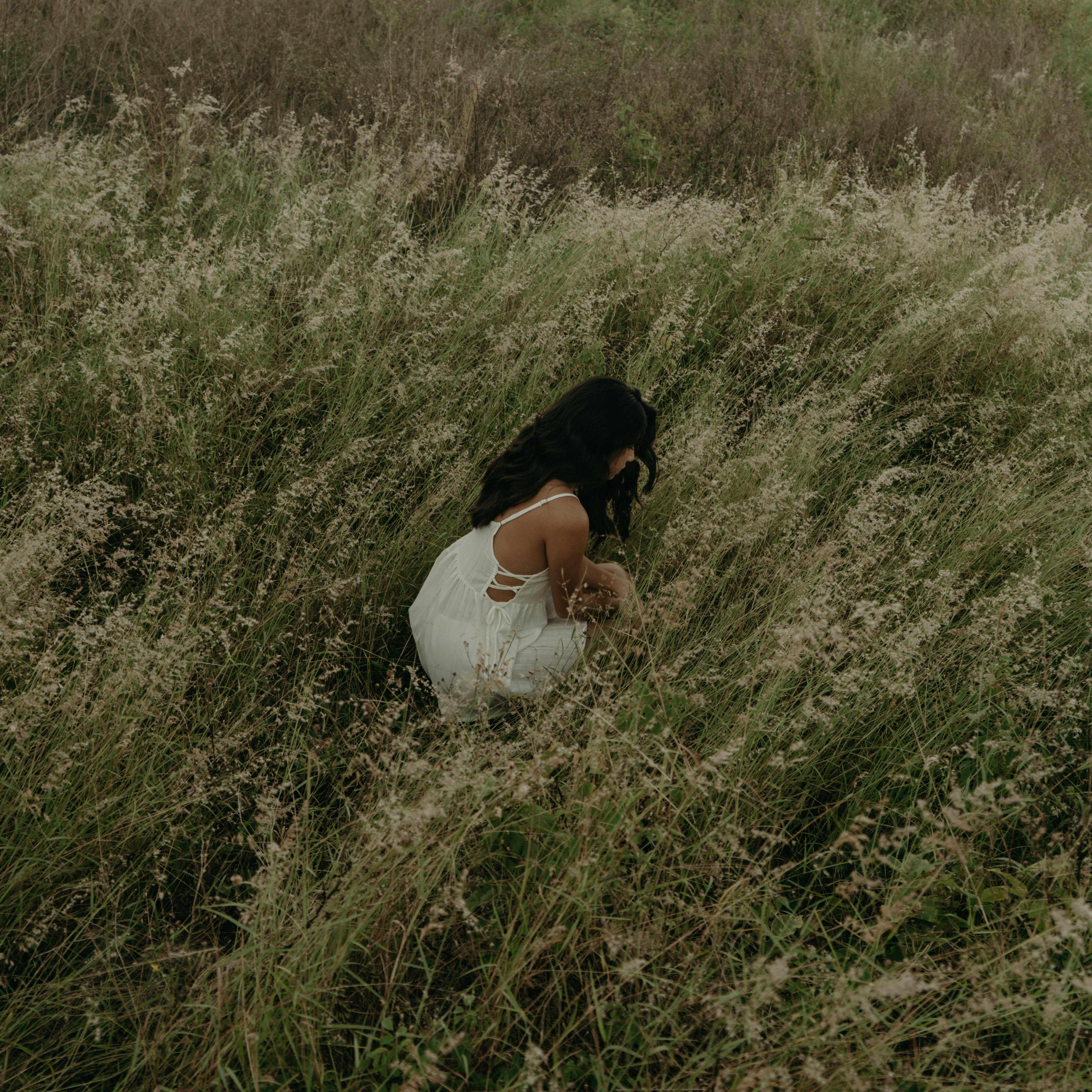 A woman crouches in a grassy rural field, wearing a white dress.