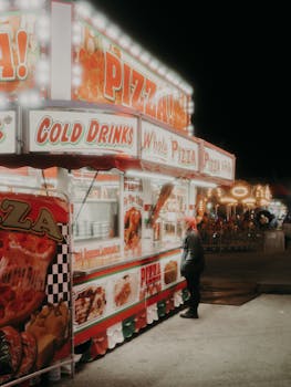 Illuminated pizza and drinks stall at night carnival with a nostalgic vibe.