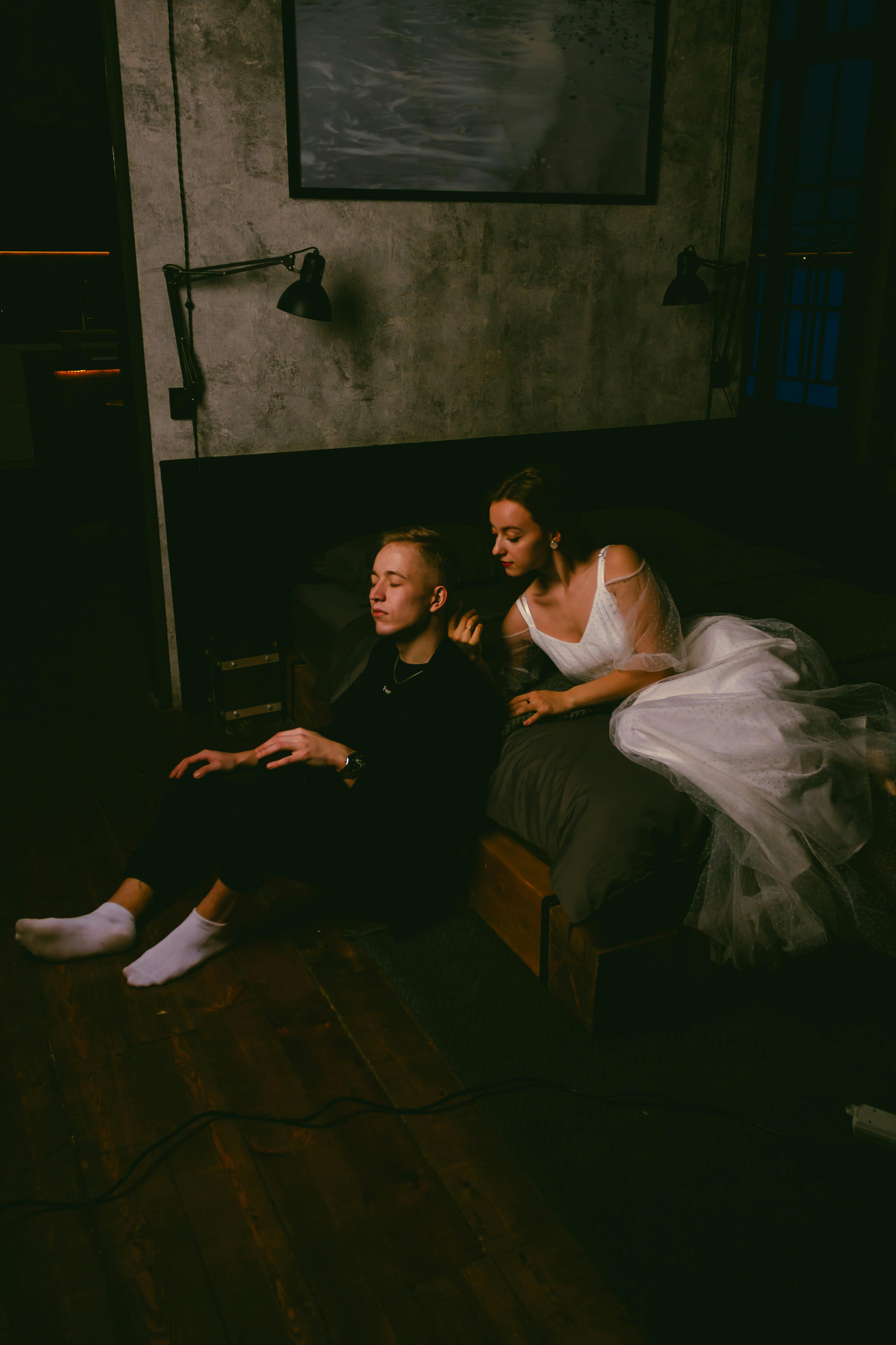 Couple sitting on bed in dimly lit bedroom, capturing an intimate and relaxed moment.