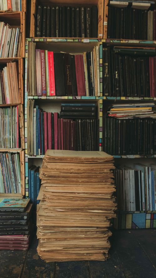 Free A tall stack of books in front of filled, colorful bookshelves, creating a cozy library feel. Stock Photo