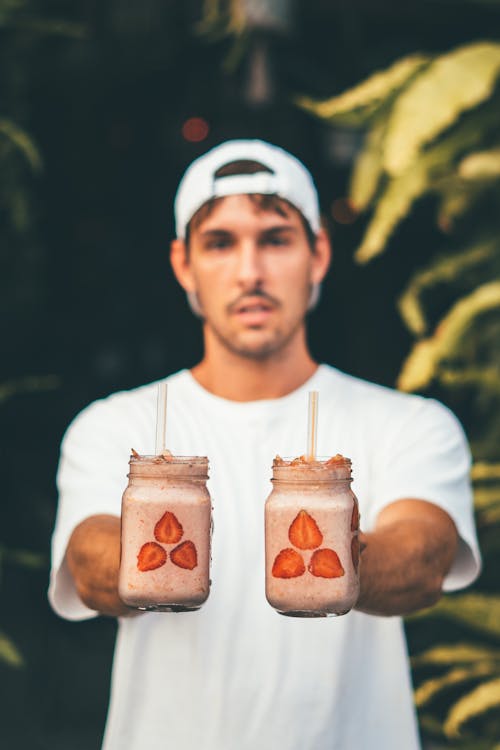 Man Wearing White Crew-neck T-shirt and Fitted Cap Holding Two Glass Jars