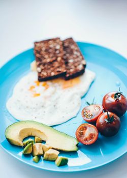 Delicious and nutritious breakfast of fried eggs, avocado, and cherry tomatoes on a blue plate.