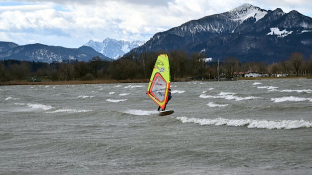A windsurfer rides waves with stunning mountains in the background on a cloudy day.