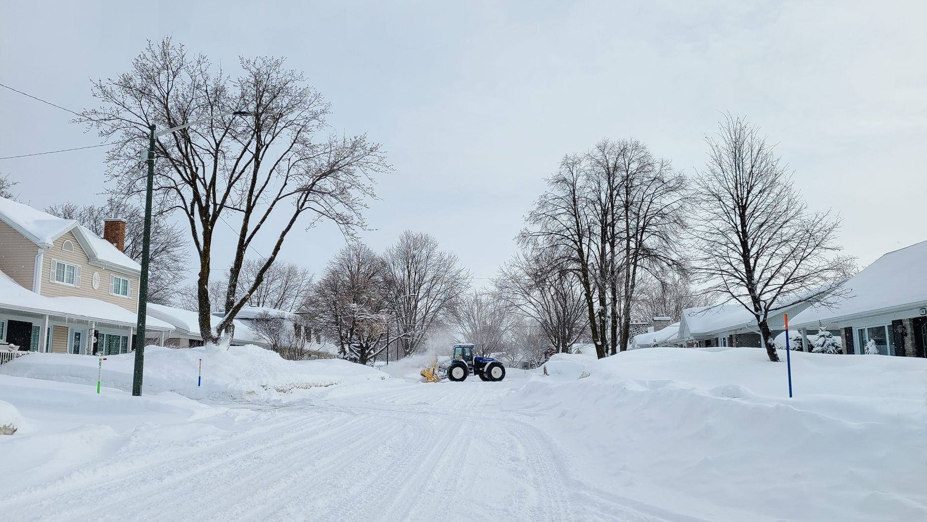 Tractor clearing snow in a wintery neighborhood in Quebec. Leafless trees line the snow-covered street.