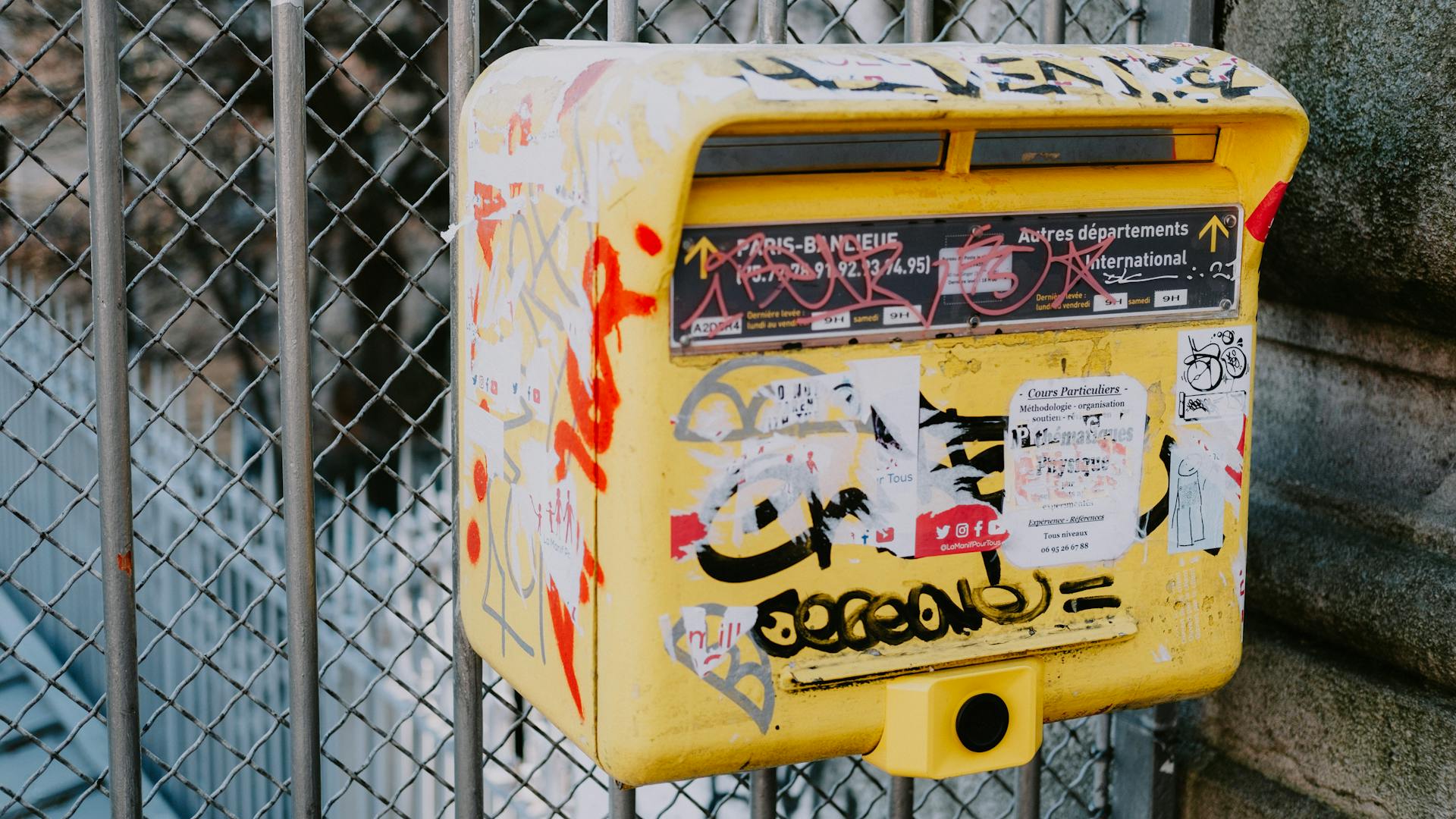 A yellow mailbox with graffiti and stickers in an urban environment, showcasing vibrant street art.