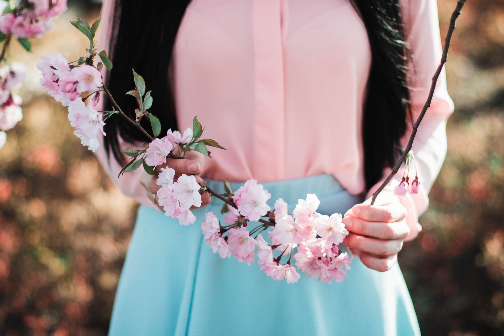 Woman holding pink cherry blossoms in spring. Fashionable attire adds to the serene outdoor scene.