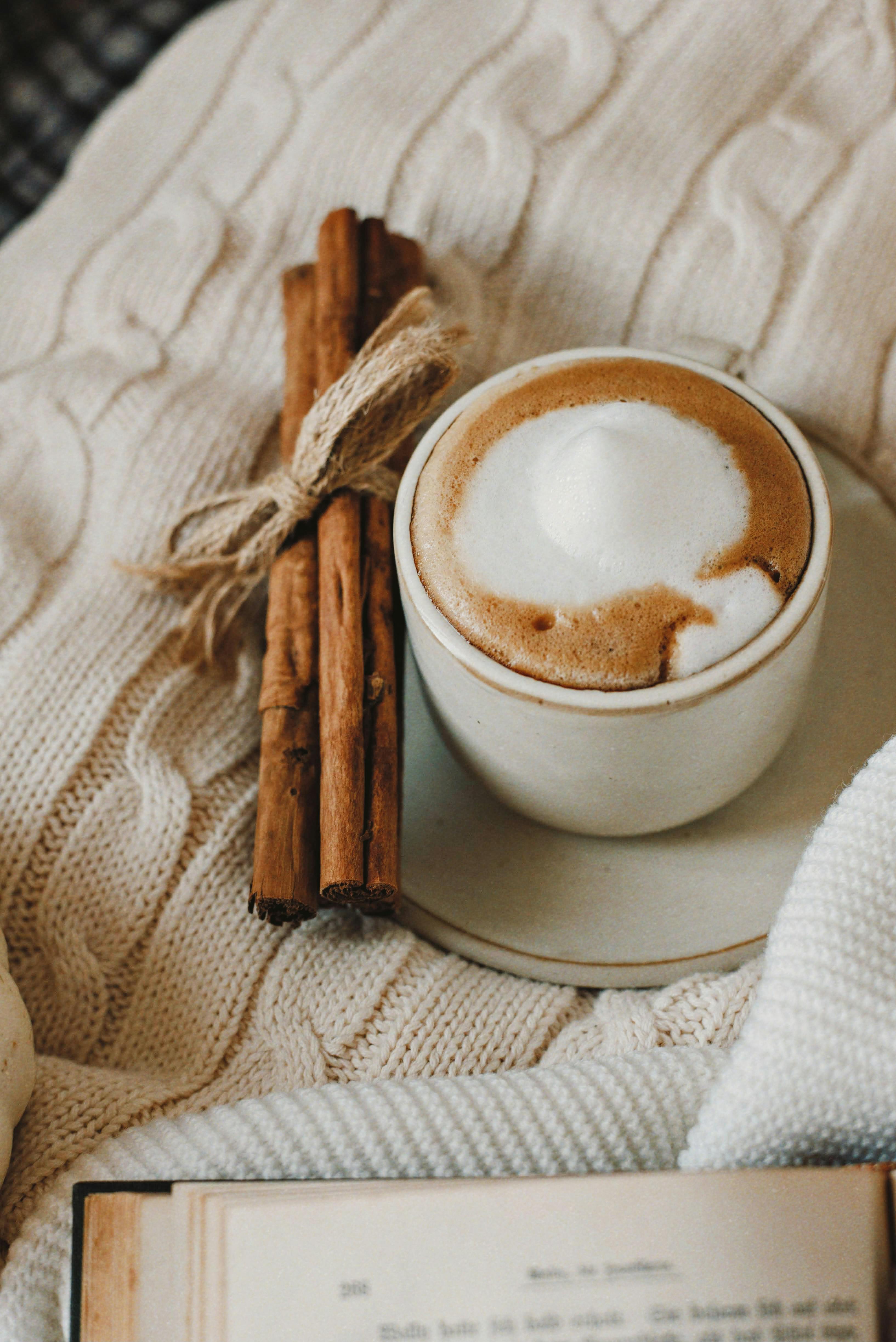 A warm cappuccino with cinnamon sticks on a knitted blanket evokes cozy vibes.