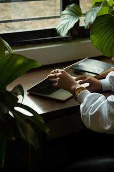 A woman working remotely on a laptop by a window, surrounded by lush houseplants.