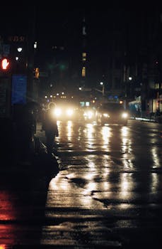 Moody urban night scene of a city street with headlights and reflections on wet pavement.