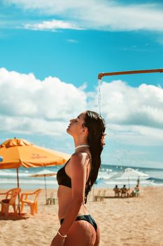 A woman in a bikini showers on a beach, embracing the sunny day and ocean waves.