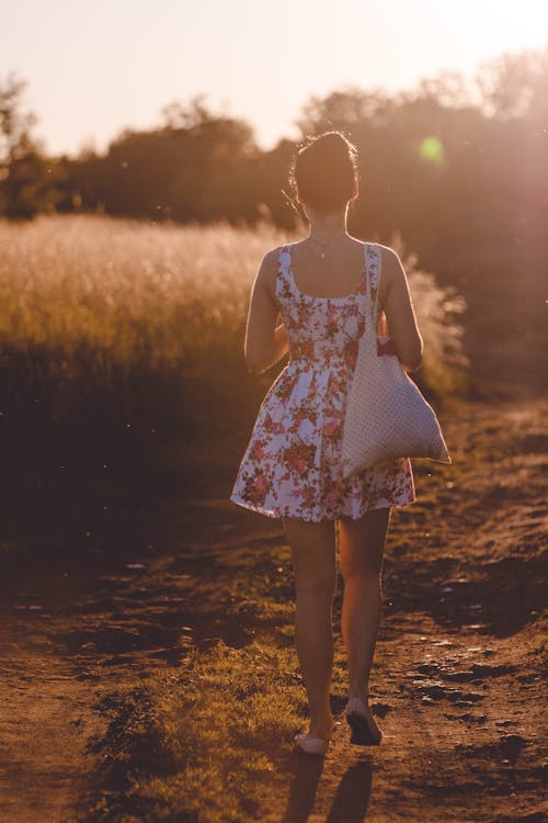 日光の下で茶色の道を歩く白とピンクの花のノースリーブドレスの女性
